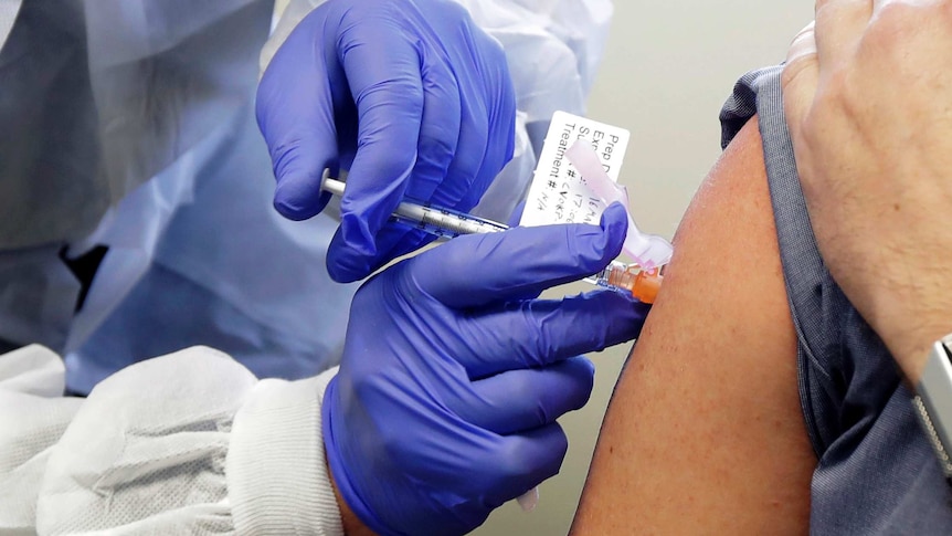 A pair of hands in latex gloves holds a vaccine vial and syringe as it is injected into an arm