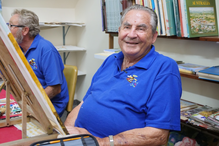 An older man with grey hear sits in front of a canvas smiling.