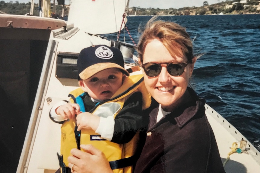 A woman in sunglasses holds a baby in a yellow life jacket, on a boat