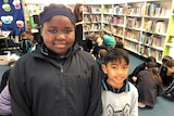 Students Sohaiba Mustafa and Christian Buelva standing in the school library with other students and books in the background.