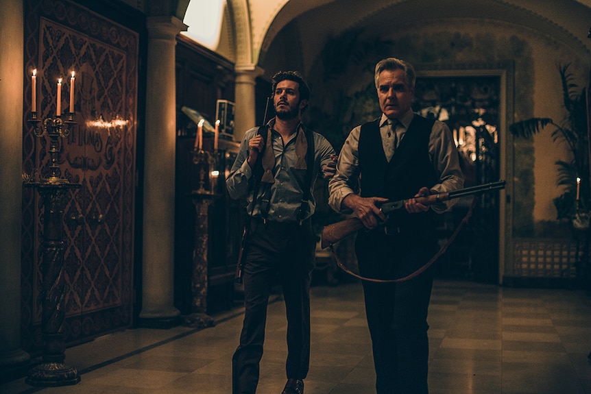 Adam Brody and Henry Czerny wear shirt and suit pants with firearms while walking down candle lit ornate Gothic style hallway.
