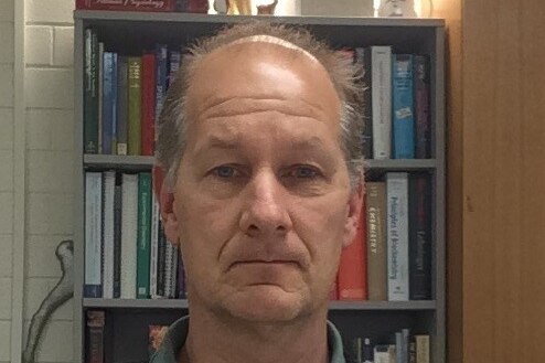 A middle-aged man stands in front of a bookcase with a stern facial expression.