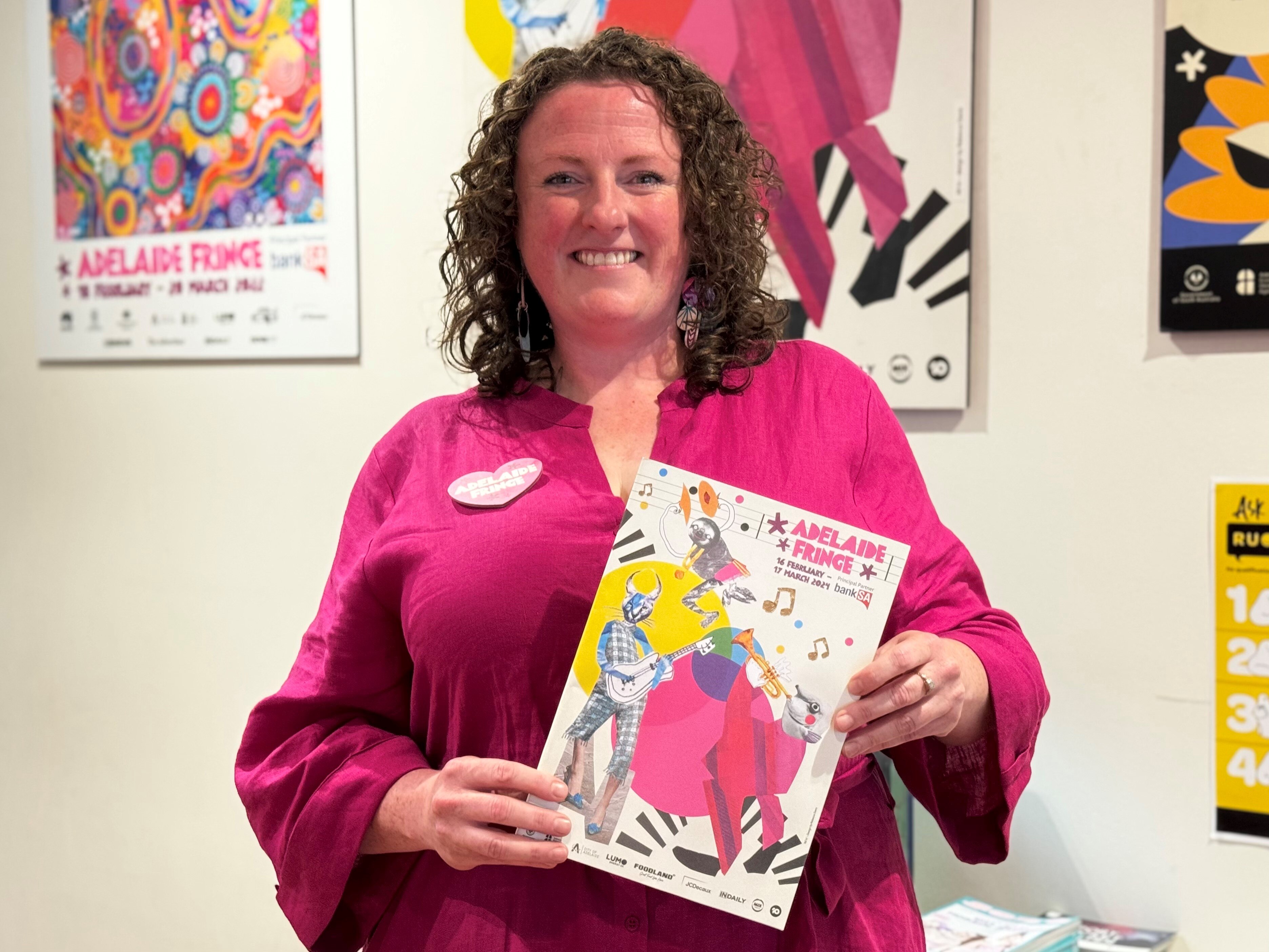 A woman smiling and holding up the Adelaide Fringe guide book in front of a wall of posters