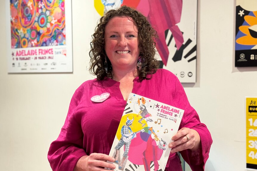 A woman smiling and holding up the Adelaide Fringe guide book in front of a wall of posters