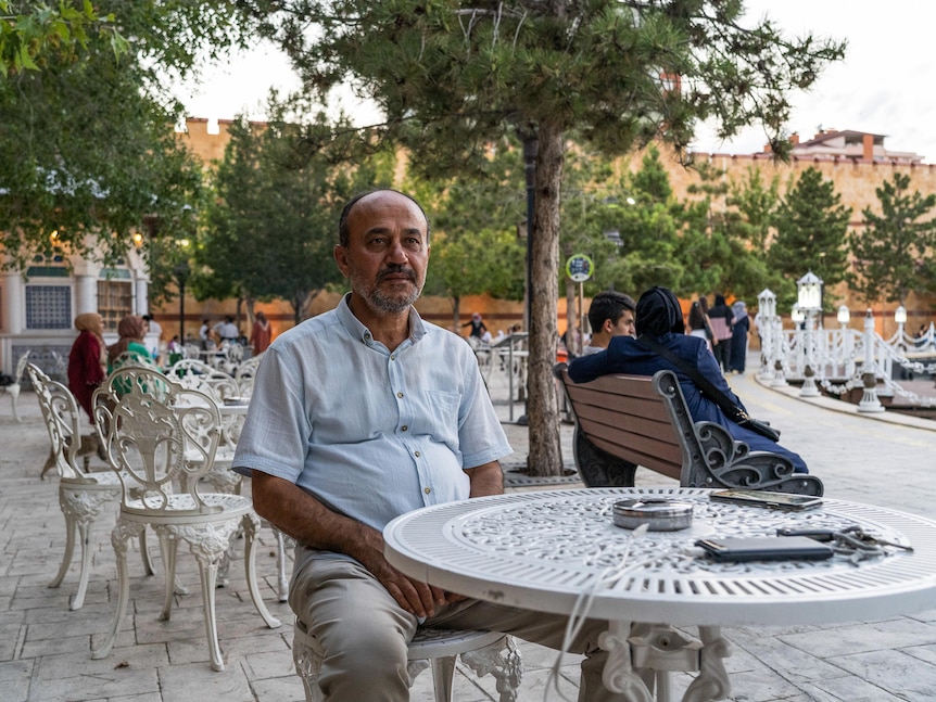 A middle-aged man sits at a white patio table in a peaceful courtyard. Others behind him sit at park benches