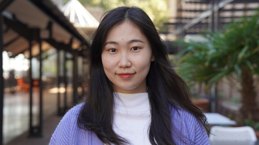 A woman in purple and white top stands on a uni campus with a smile.