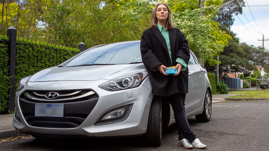 A woman leans on a small silver hatchback holding medication boxes