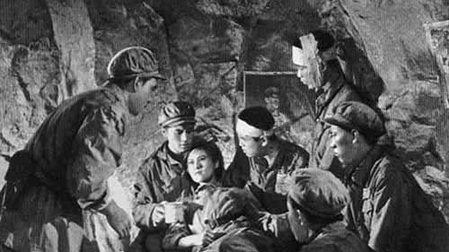A black and white image shows a woman lying own among men tending to her care in an underground military base.