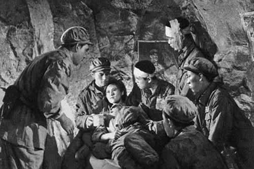 A black and white image shows a woman lying own among men tending to her care in an underground military base.