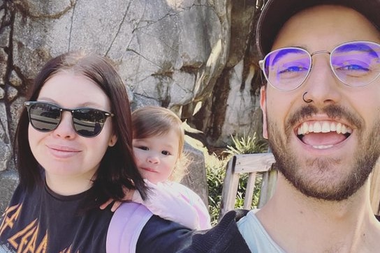 A couple holding a toddler smile at the camera.