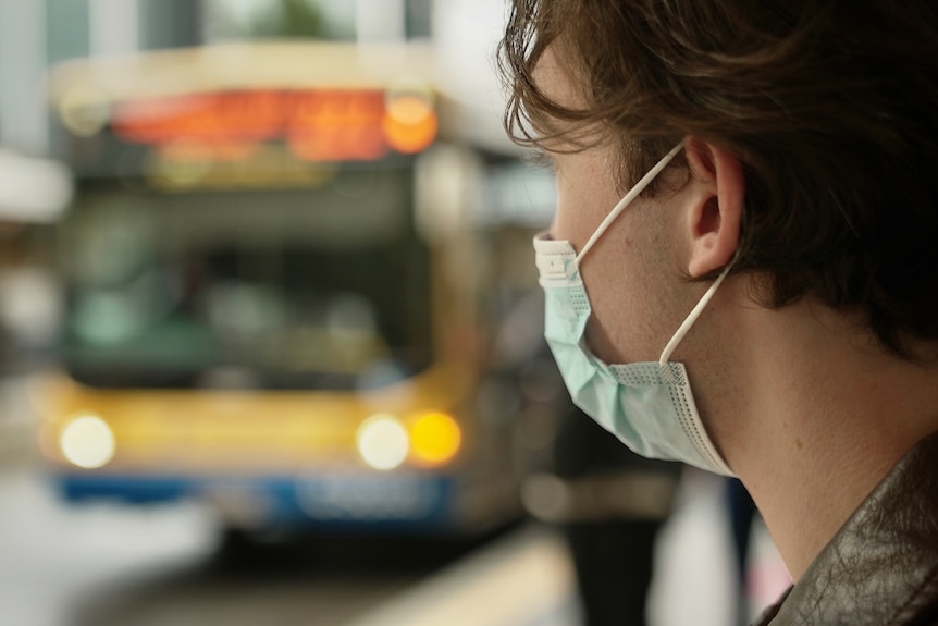Man wearing face mask sitting at Brisbane bus stop, bus blurred in the background.