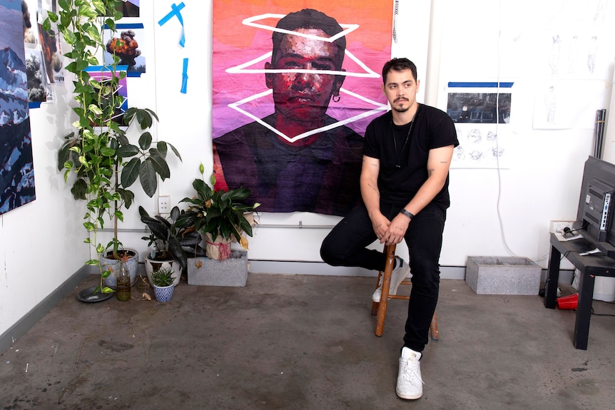 A man sits on a stool in front of a large painting. He is looking thoughtful.  