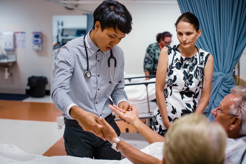 A young man with a stethoscope holds the hands of a patient while a woman with a stethoscope looks on.