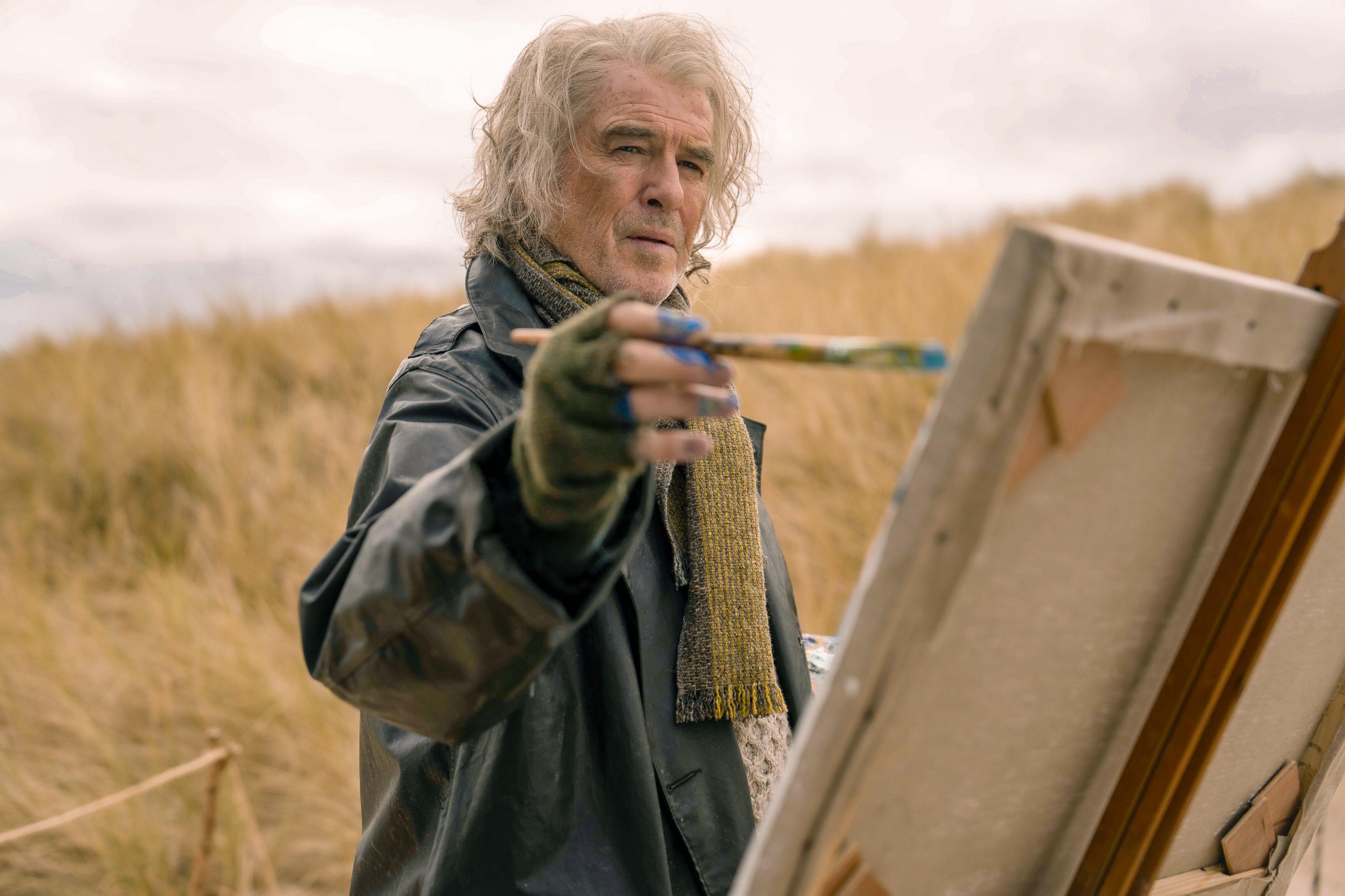 Pierce holds a paintbrush up to a canvas while standing in a brown grassy field on an overcast day.