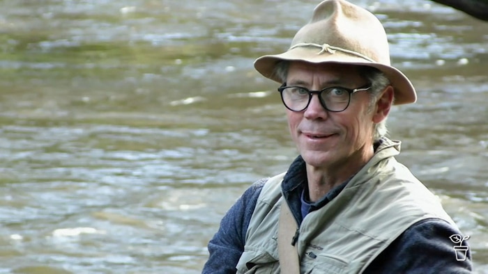 Man wearing a hat and fishing vest wading in a stream.