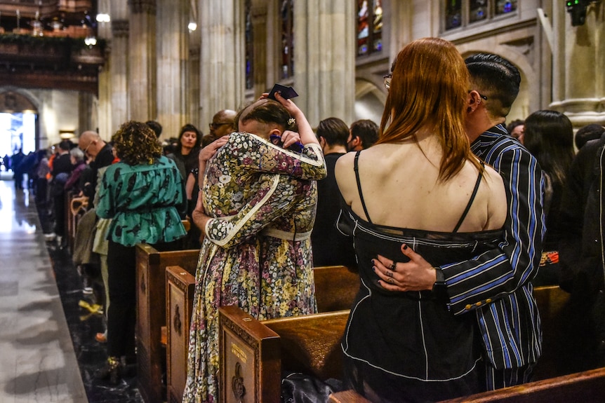 The backs of people hugging while standing in pews of a church. The church is very full.