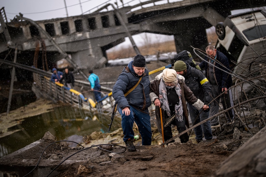 Local militiamen help an old woman crossing a bridge destroyed by artillery.