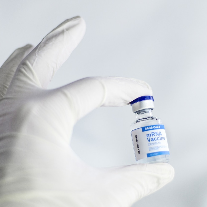 A gloved hand holds up a vial of an mRNA SARS-CoV-2 coronavirus vaccine with two fingers.
