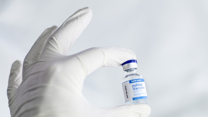 A gloved hand holds up a vial of an mRNA SARS-CoV-2 coronavirus vaccine with two fingers.