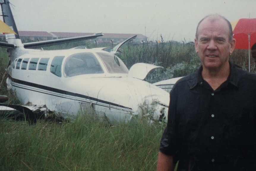 Mike Willesee stands in the rain in front of a crashed plane. People hold umbrellas in the background