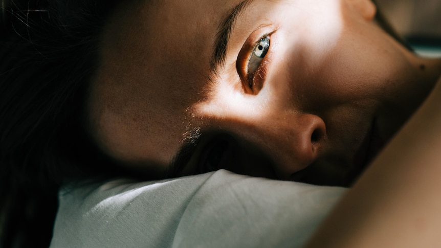 A close-up of a face lying on a pillow in the dark, with one ray of light on her eyes, which look straight ahead.