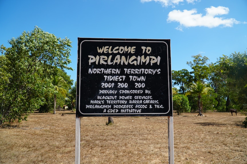 A sign reading "Welcome to Pirlangimpi'. Behind the sign is dry grass and green plants.
