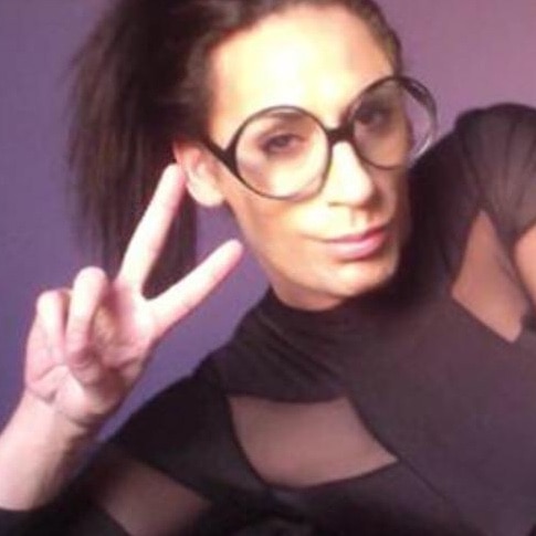Transgender sex worker CJ Palmer, who is wearing glasses and has a ponytail, makes a peace sign at the camera.