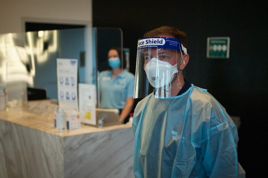 A man wearing full PPE and a woman in the background behind a screen.