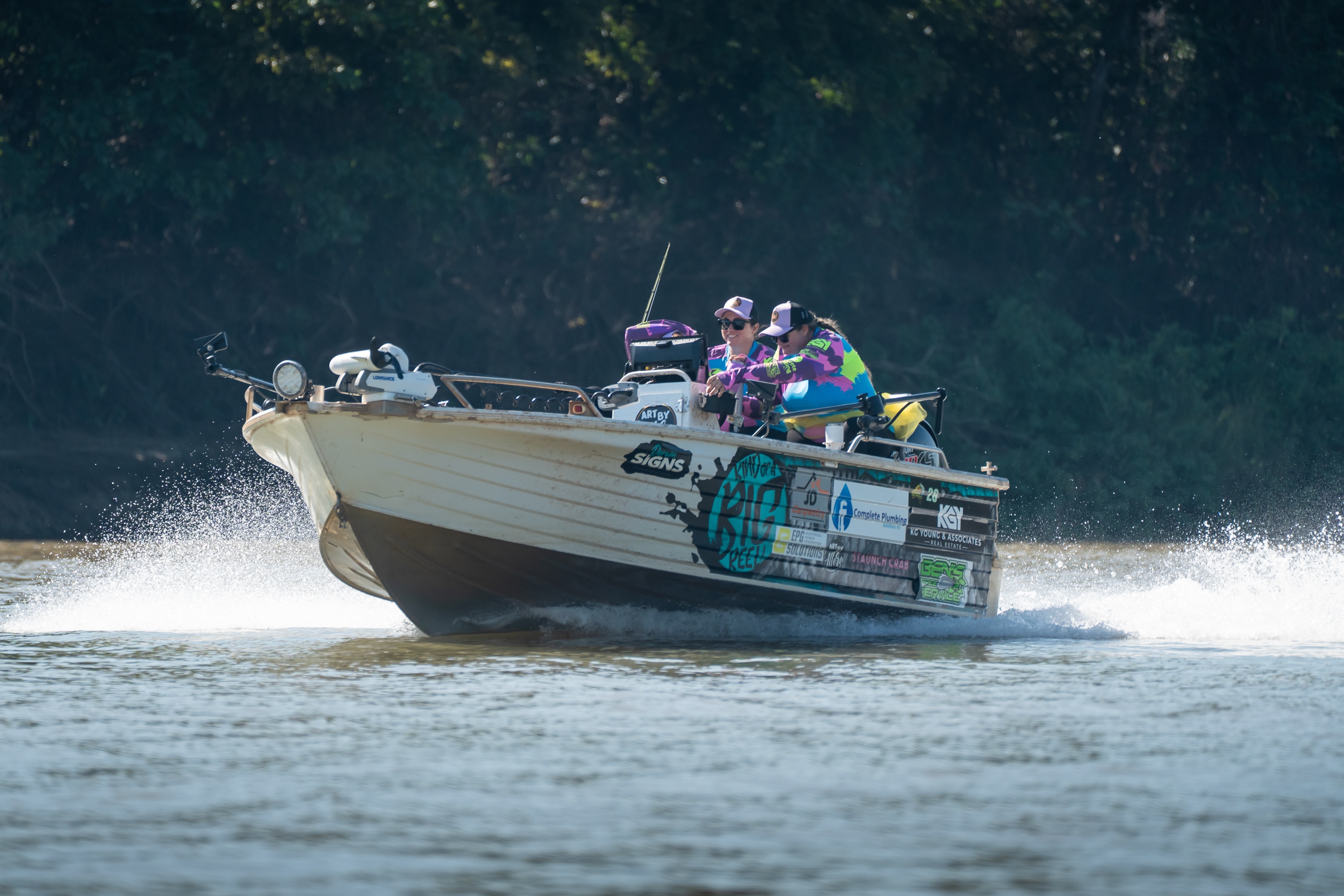 A white small fishing boat cruising along the water, two women in purple / green long sleeve tops driving it.