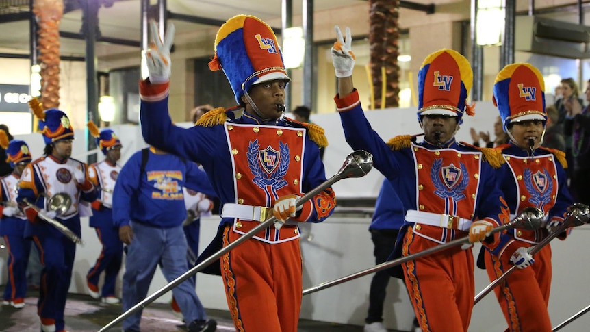 Three black men wearing elaborate marching costumes, walking along and showing the peace sign with their hands. 