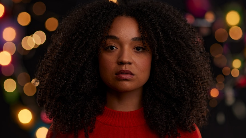 A Black woman in her late 20s with curly hair and red top, bright lights in the night behind her