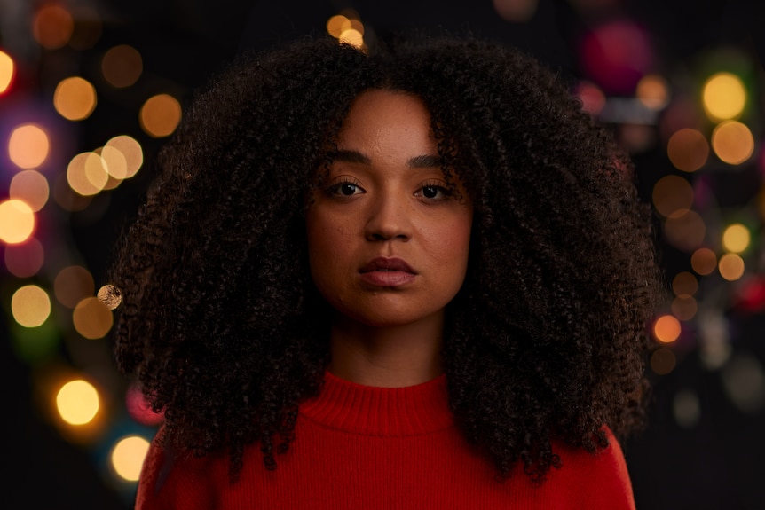 A Black woman in her late 20s with curly hair and red top, bright lights in the night behind her