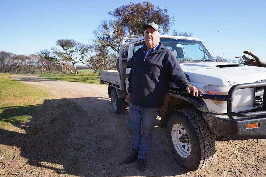A man in a navy jumper stands next to a ute on a curving dirt road.