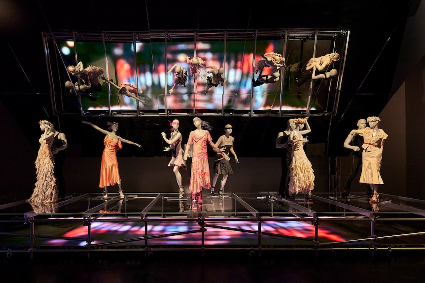 Nine mannequins positioned to appear in-motion wear intricate garments on a platform with a large mirror hanging overhead.