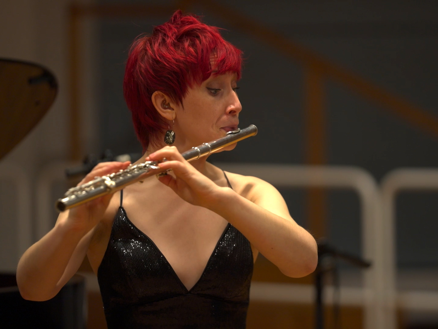 Eliza Shephard plays the silver flute in a wooden concert hall. She has short red hair and wears a shiny black top.