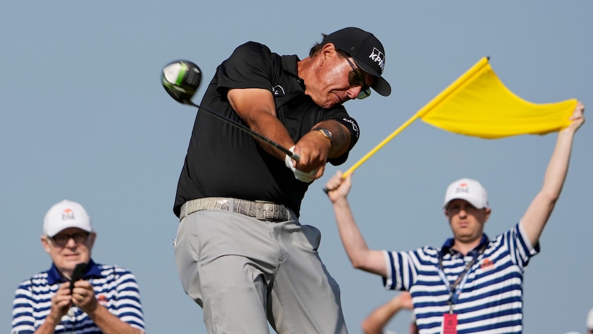 Phil Michelson grimaces as he hits a shot, with a man holding a yellow flag stands in the background