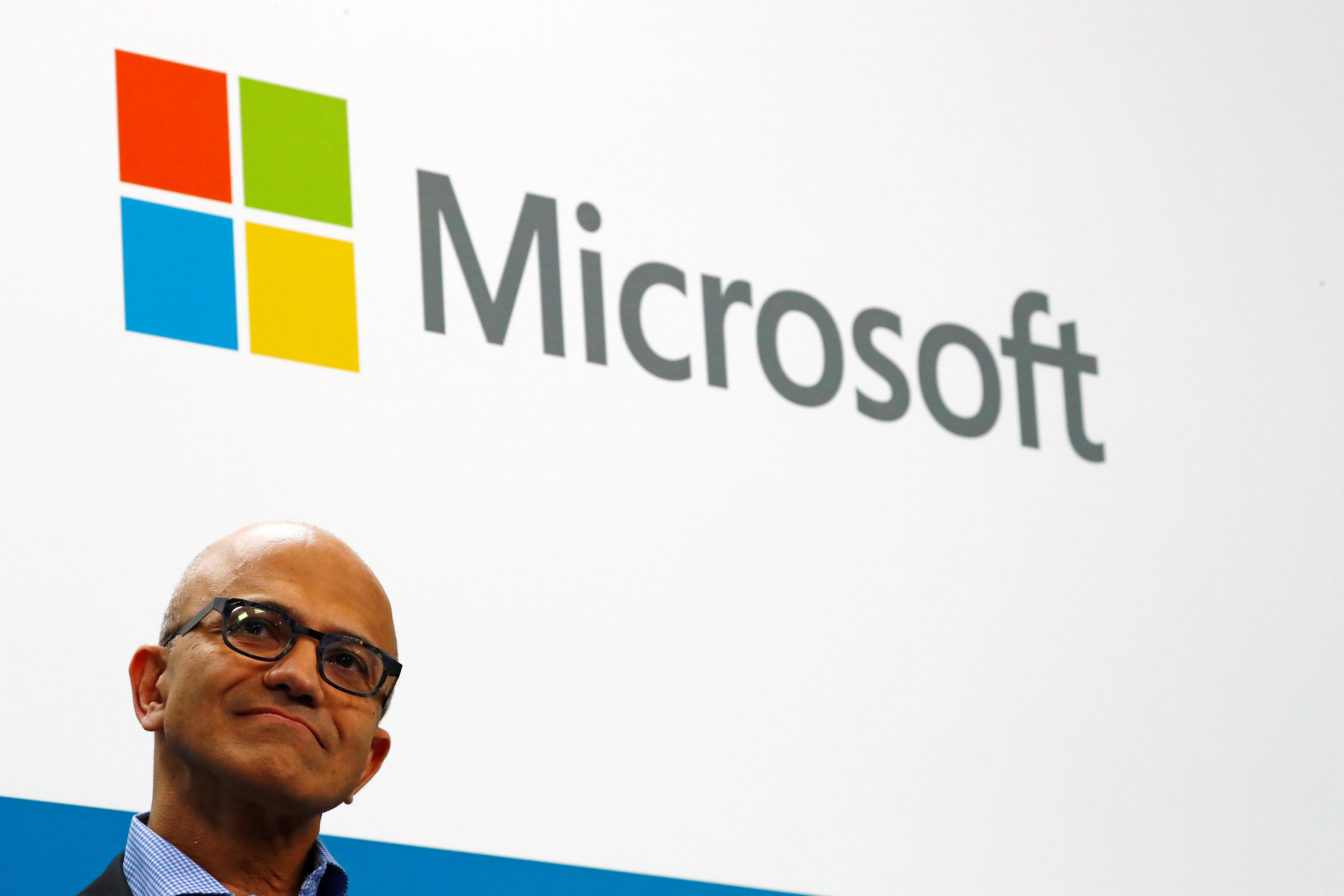 The face of Microsoft CEO Satya Nadella, wearing glasses, frowning slightly in front of the Microsoft logo behind him.