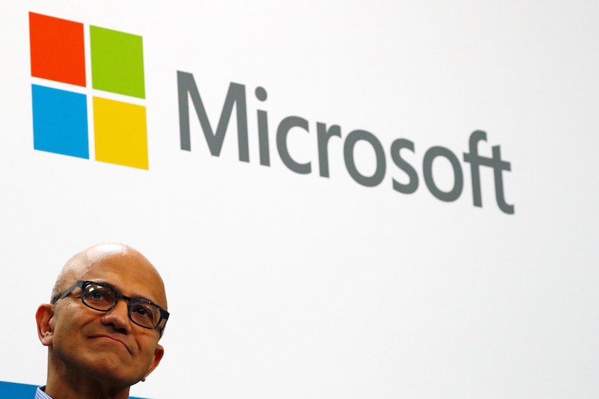 The face of Microsoft CEO Satya Nadella, wearing glasses, frowning slightly in front of the Microsoft logo behind him.