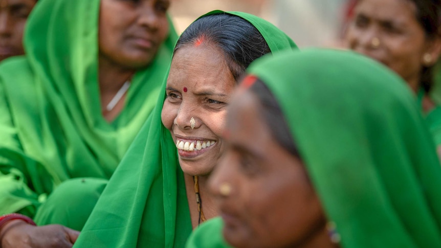 A woman smiles while sitting among a group of women all dressed in green saris.