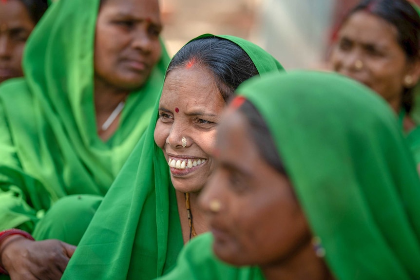 A woman smiles while sitting among a group of women all dressed in green saris.