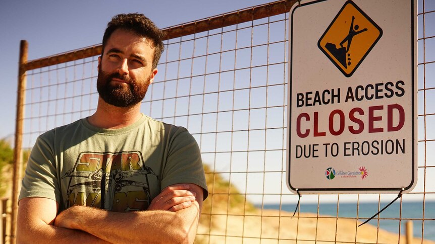 A man in a khaki t-shirt stands at a beach with arms folded next to a fence with a sign that says "beach closed due to erosion".