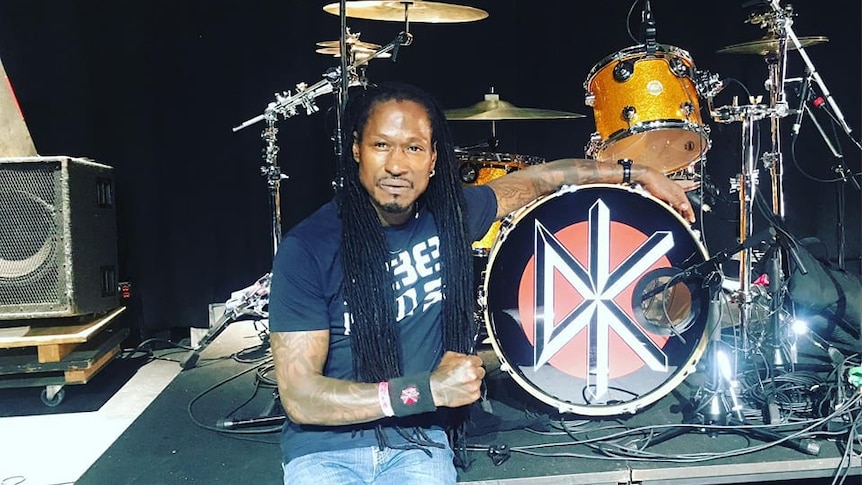 D.H. Peligro sits next to a drum kit with Dead Kennedys logo on stage at the Ventura Theater clenching a fist.