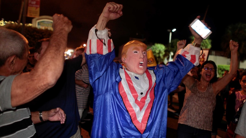 A man wearing a donald trump mask and outfit rejoices among a crowd of people in the streets of Miami