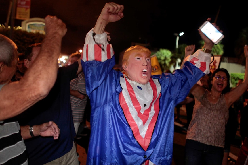 A man wearing a donald trump mask and outfit rejoices among a crowd of people in the streets of Miami