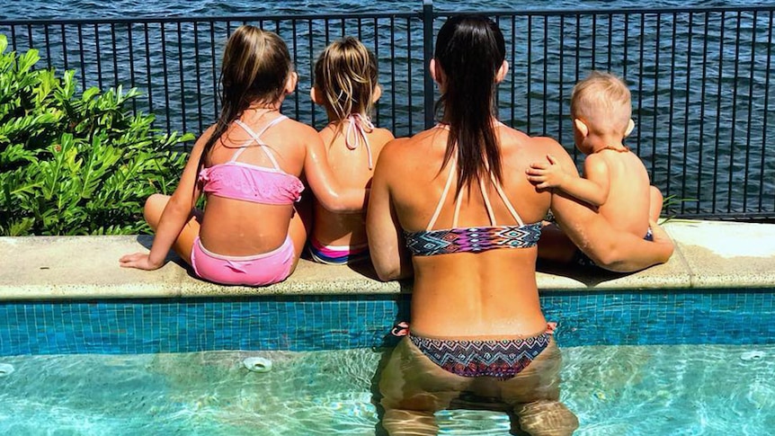 A woman half immersed in a swimming pool with three small children, all of them staring out at water with backs turned