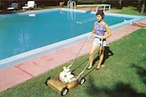 A woman in the 1960s pushing a lawnmower beside a pool.