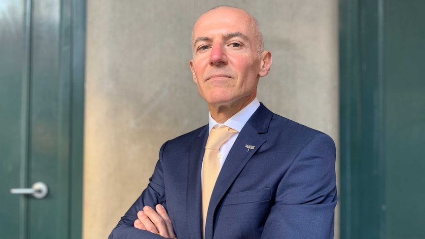 Silvio Del Vecchio stands with his arms folded wearing a blue suit and pale yellow tie