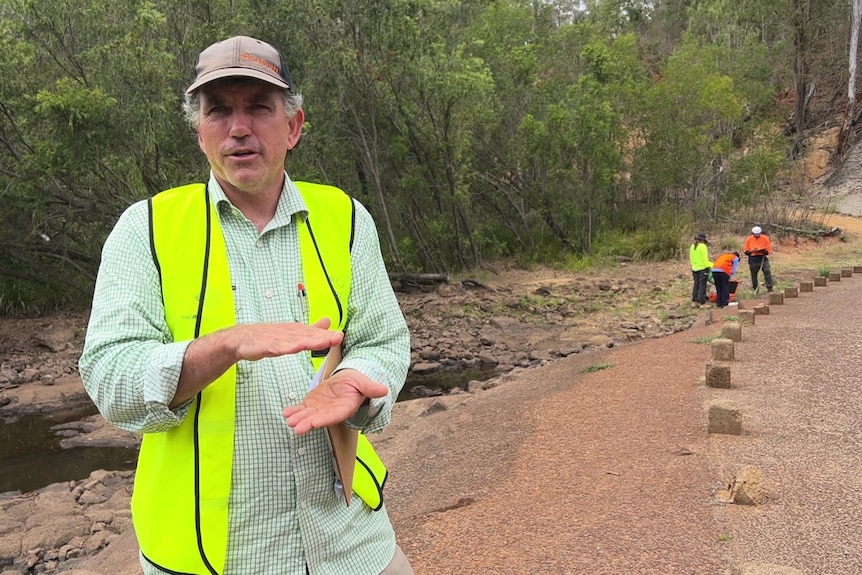 A man in a high-vis vest and cap stands along a road causeway with a waterway behind 