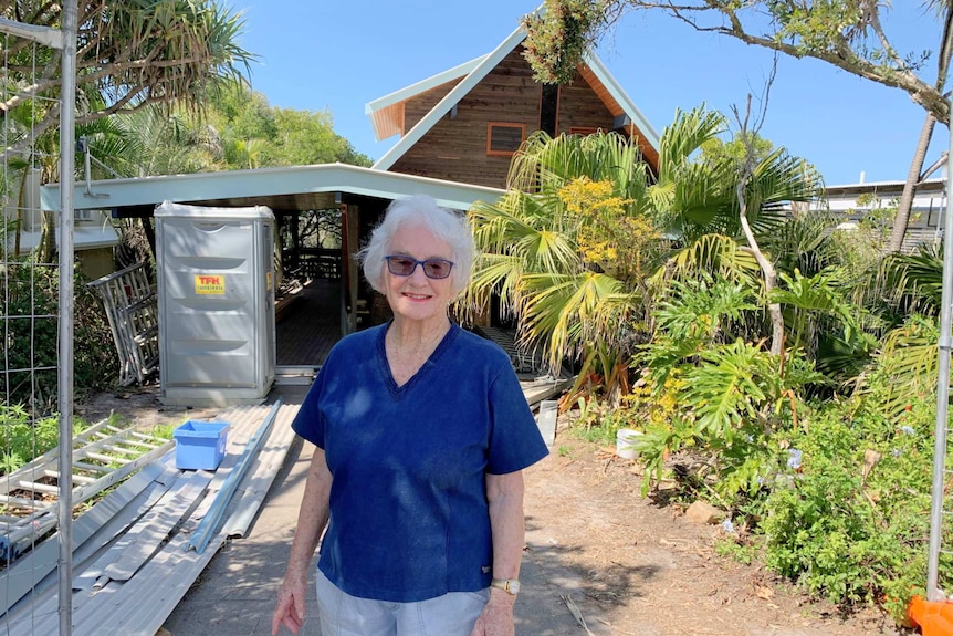 Woman with grey hair stands in front of an A-frame house under construction. She is smiling.