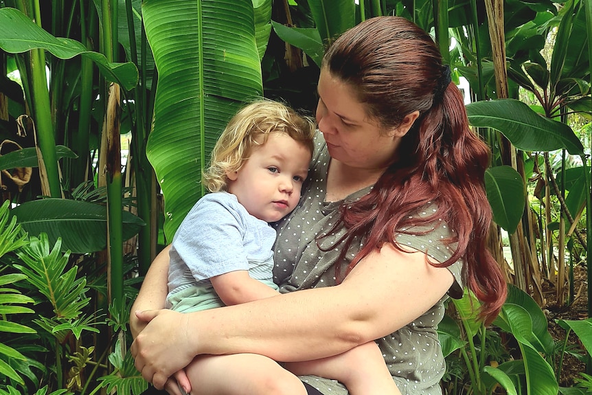 Nataasha Torzsa holds her young son. She's standing in front of tropical plants.